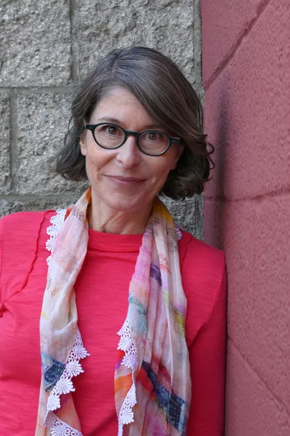 Jennifer looks at us with a closed-mouth smile. Leaning against a brick wall, she stands in a bright coral top with a scarf draped around her neck.
